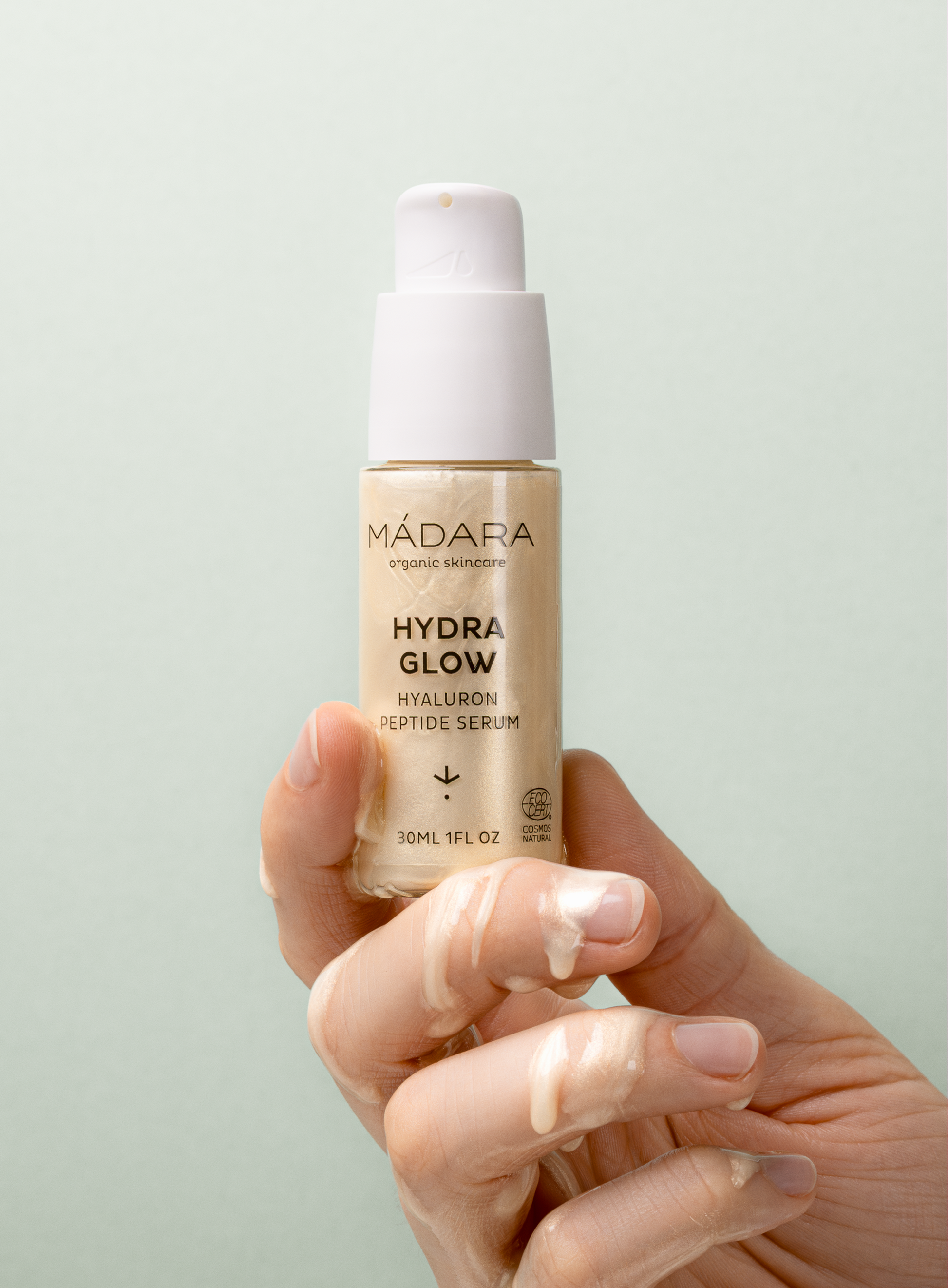 Hand holding MÁDARA Hydra Glow Hyaluron Peptide Serum 30ml bottle with serum texture on fingers, highlighting hydrating and glowing skincare formula
